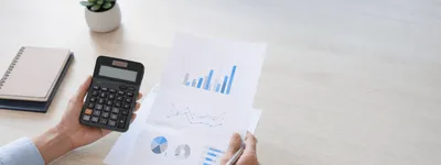 Mãos segurando uma calculadora e uma caneta sobre folhas com gráficos de barras, linhas e pizza, calculando o retorno de um investimento