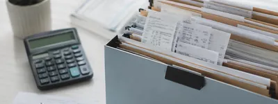 Caixa organizadora com pastas suspensas cheias de recibos e comprovantes, calculadora e caneta sobre papéis ao lado, numa mesa de escritório