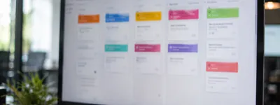 Monitor exibindo um quadro kanban com colunas coloridas e cartões de tarefas, sobre uma mesa de escritório com planta ao lado