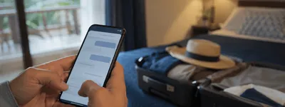 Hóspede enviando mensagem pelo celular em quarto de hospedagem, com mala e chapéu de viagem ao fundo