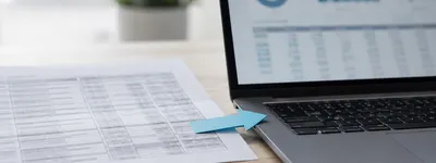 Seta de papel azul apontando de uma planilha impressa para um notebook aberto exibindo gráficos e dashboard, sobre mesa de madeira com planta ao fundo