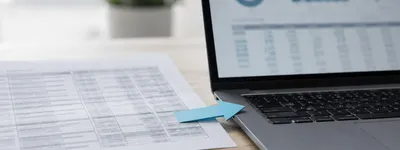 Seta de papel azul apontando de uma planilha impressa para um notebook aberto exibindo gráficos e tabelas na tela