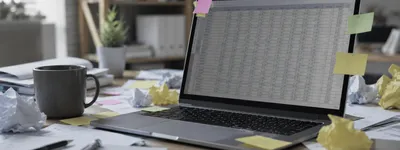 Notebook aberto mostrando planilha do Excel cercada de post-its e papéis amassados sobre a mesa