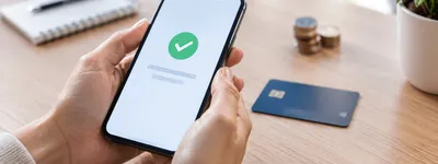 Mãos segurando um celular exibindo uma tela genérica de confirmação de pagamento, com um cartão de crédito desfocado ao lado sobre uma mesa de madeira clara e moedas fora de foco ao fundo