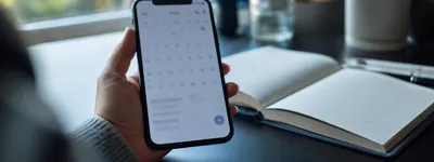 Close de mãos segurando um celular exibindo um app de calendário mensal, com um caderno aberto e vazio ao fundo sobre uma mesa escura