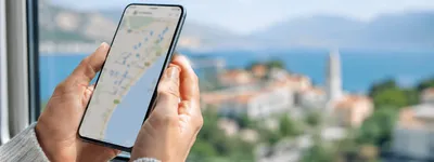 Mãos segurando um celular com um mapa aberto na tela, em frente a uma janela com vista para uma cidade litorânea com casario e mar ao fundo