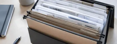 Caixa organizadora preta com pastas suspensas cheias de documentos, comprovantes e recibos, sobre uma mesa de escritório