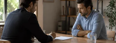 Dois homens sentados à mesa numa negociação, um deles com os braços cruzados e expressão desconfiada, enquanto o outro assina um documento
