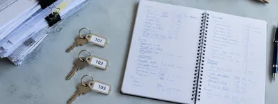 Caderno espiral aberto com anotações manuscritas em tabela, ao lado de três chaves com etiquetas numeradas 101, 102 e 103 e uma pilha de papéis presos com clipes
