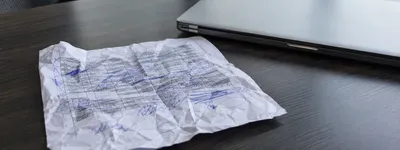 Folha impressa de planilha amassada e rabiscada com caneta azul, jogada sobre mesa escura ao lado de um laptop fechado