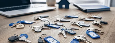 Várias chaves numeradas de imóveis espalhadas sobre a mesa, ao lado de notebook e miniatura de casa