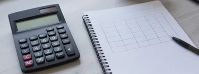 Calculadora preta ao lado de um caderno de espiral aberto em página com tabela desenhada à mão e uma caneta apoiada sobre o papel, xícara desfocada ao fundo