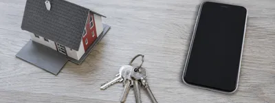 Uma casa em miniatura, um molho de chaves e um celular dispostos lado a lado sobre uma mesa de madeira