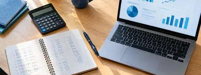 Calculadora, caderno com anotações de valores e notebook com gráficos sobre mesa de escritório em luz natural