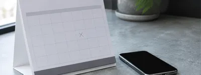 Calendário de mesa com um dia marcado por um X, ao lado de um celular com a tela apagada sobre a bancada