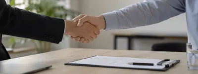 Aperto de mãos entre duas pessoas sobre uma mesa com um contrato numa prancheta ao lado