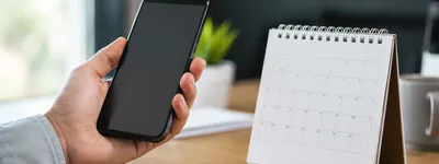 Mão segurando um celular com a tela apagada ao lado de um calendário de mesa espiral em branco sobre uma mesa de madeira