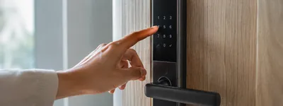 Close de uma mão digitando um código no teclado numérico de uma fechadura eletrônica preta instalada em porta de madeira