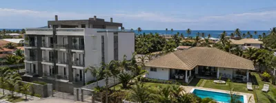 Vista de bairro litorâneo com prédio de studios compactos ao lado de casa ampla com piscina, em dia ensolarado