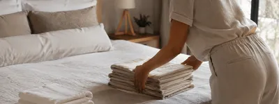 Mulher arrumando uma pilha de lençóis dobrados sobre a cama de um quarto de temporada, com toalhas dobradas ao lado