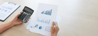 Mãos segurando uma calculadora e folhas impressas com gráficos de barras, linhas e pizza sobre a mesa